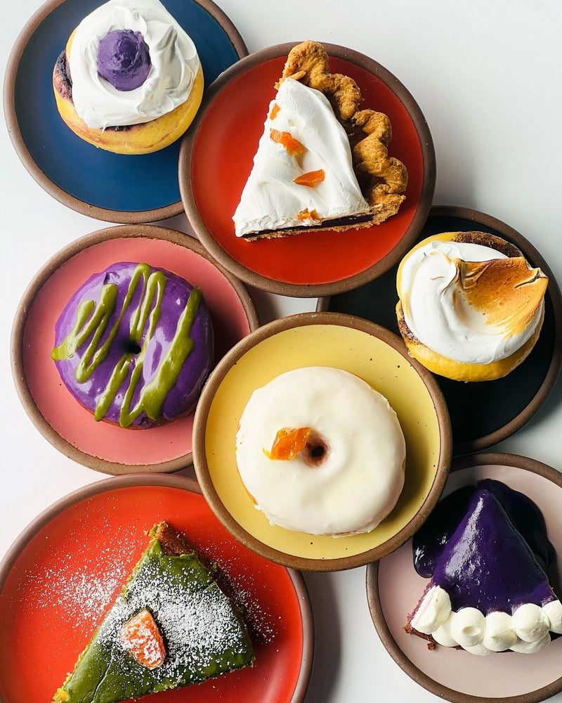 An artful overhead view of different desserts (donuts, cupcakes, pie slices) each on different colored ceramic cake plates.