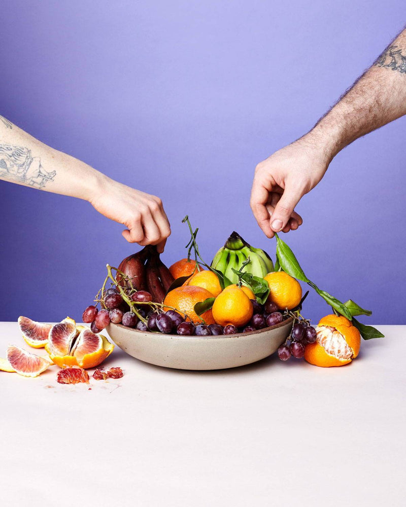 A shallow serving bowl in a warm tan brown is filled with fruit and 2 hands are reaching for it.