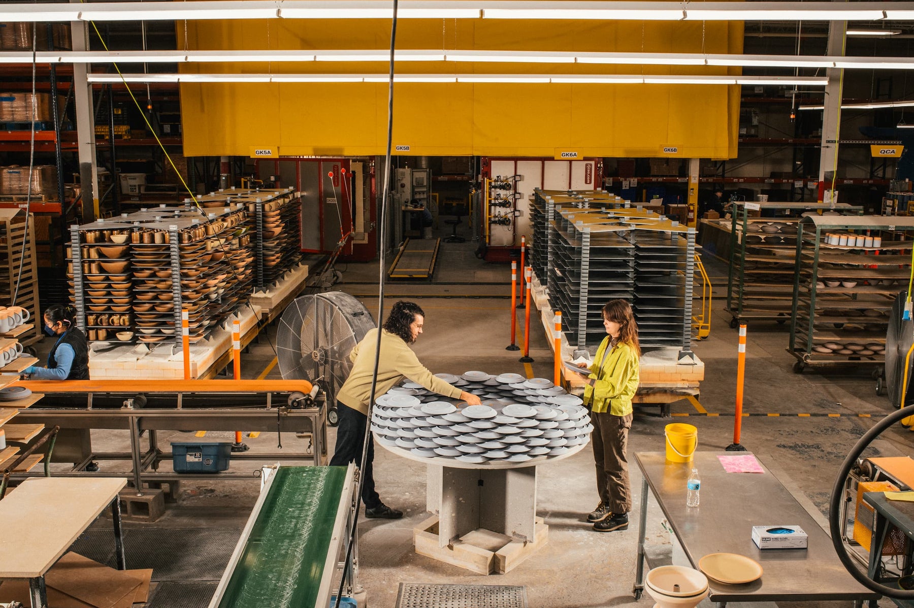 A ceramic pottery factory with large kiln carts filled with pots, assembly lines, and two people working in the center.