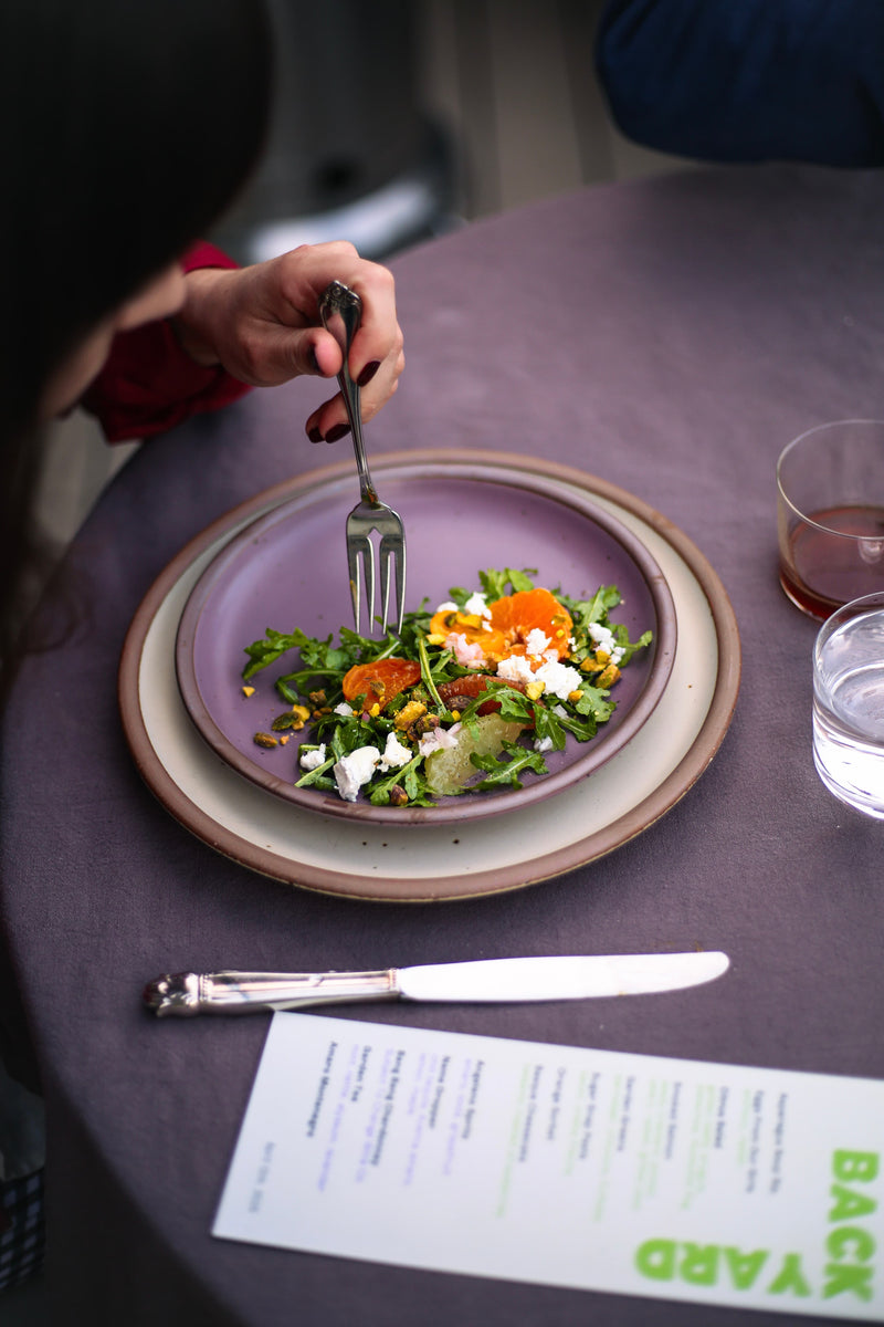 A hand holds up a fork to grab some salad from a soft mauve purple side plate next to a narrow rectangular menu.