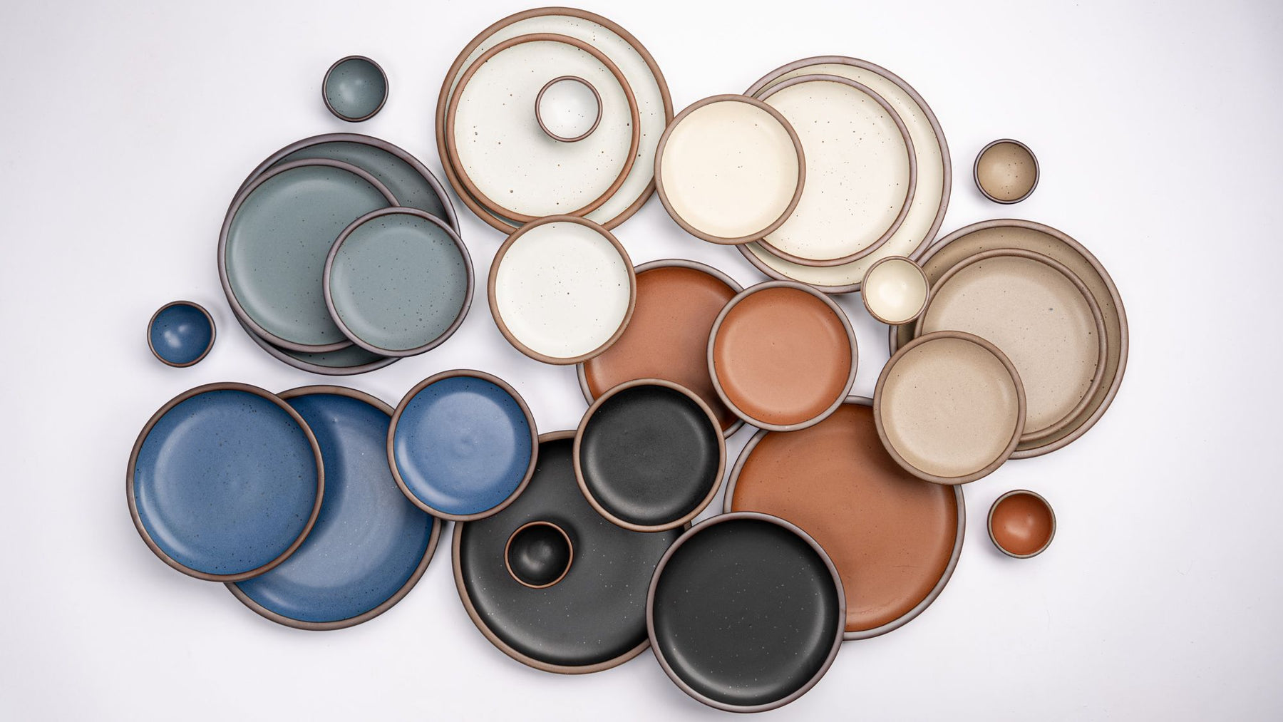 Various sizes of ceramic plates and tiny bowls in a rainbow of earthy colors are artfully laid out together against a white background.