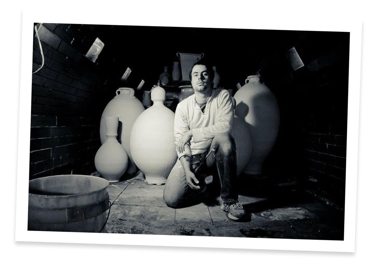 A black and white photo of Alex Matisse kneeling in front of large ceramic pieces in a wood kiln