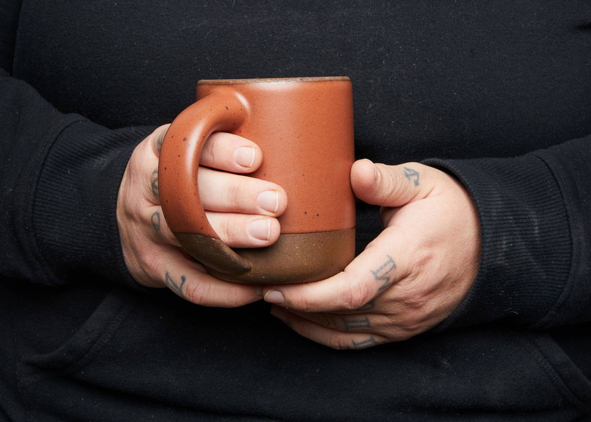 A person wearing a black sweatshirt is holding a ceramic terracotta mug with 2 hands.