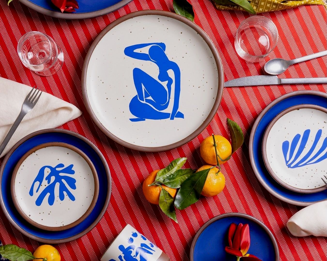 On a colorful table are plates in a bright cobalt blue, with decorated plates featuring artwork by Henri Matisse, along with silverware, glasses, and oranges.