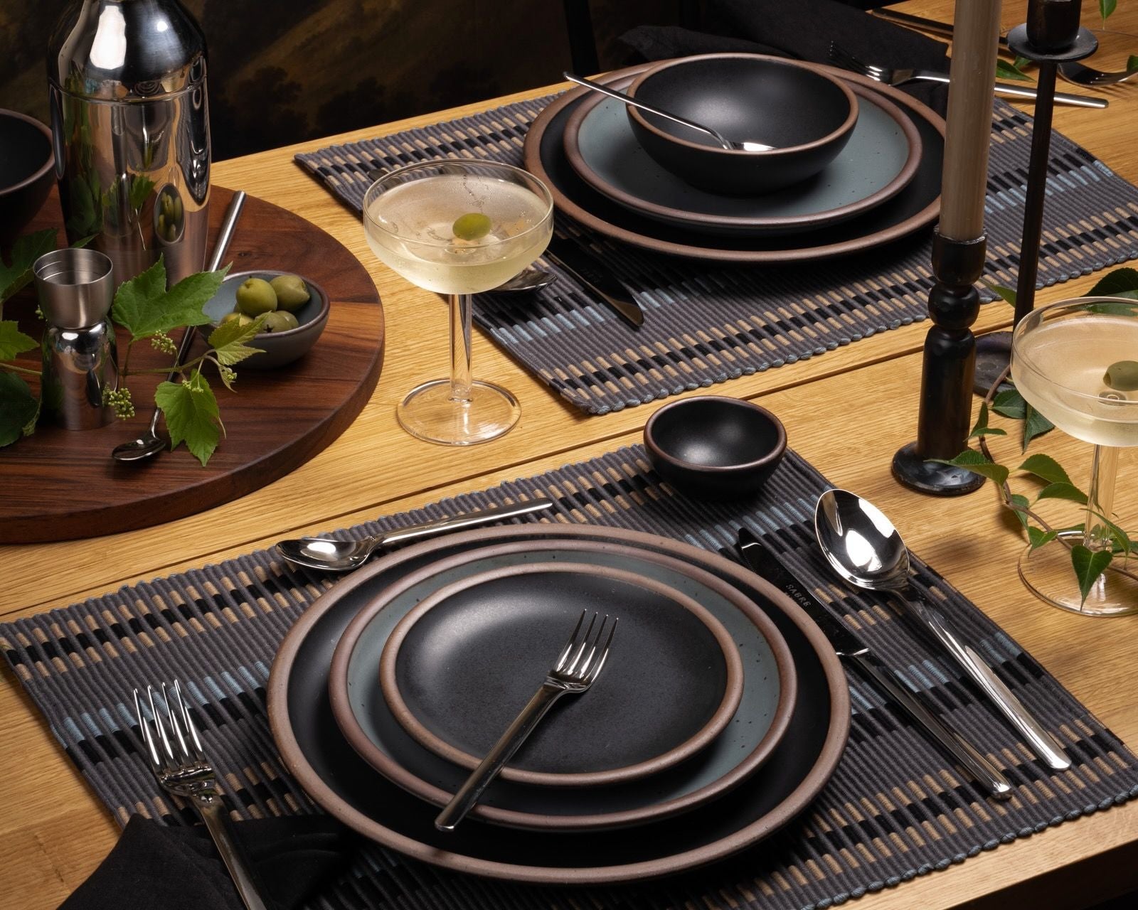 Sophisticated table setting with dark stoneware, cocktail glasses, black napkins, and stainless bar tools on a wood table with woven placemats.