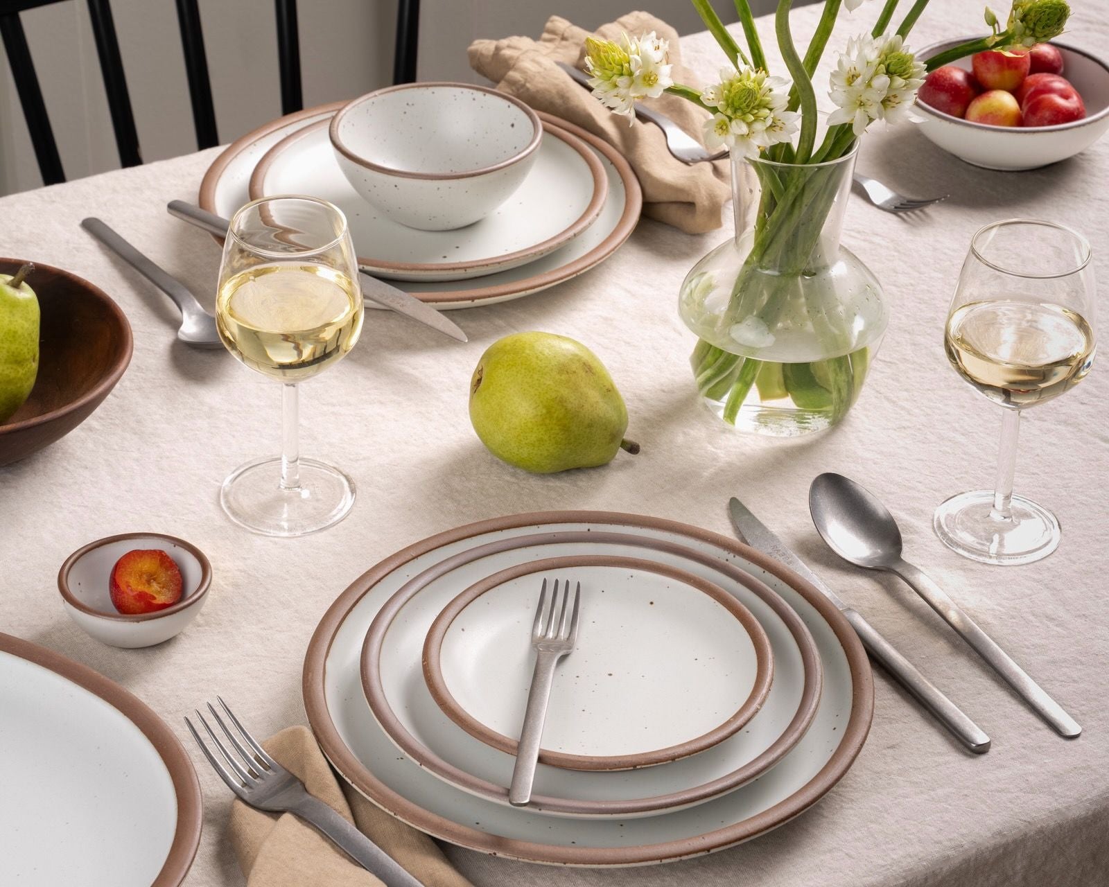 Fresh table setting with white stoneware, pears, wine glasses, and a glass vase of white flowers on a soft neutral tablecloth.
