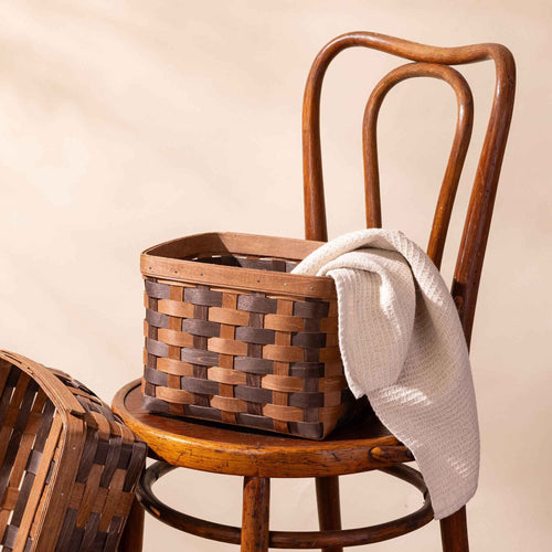Large rectangular woven wood basket in two-tone brown with an open top and reinforced rim, shown on a chair styled with a natural waffle tea towel.