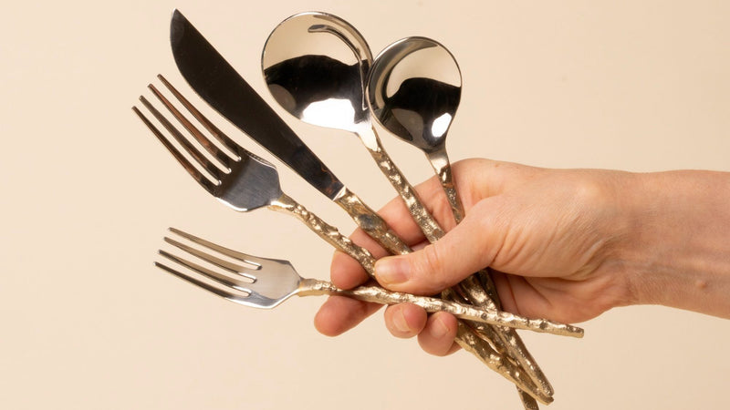A hand holds a shiny silver flatware set with hammered gold handles arranged on a beige background.