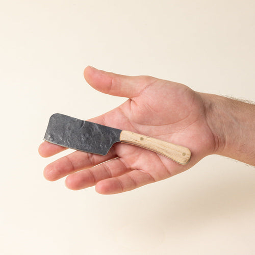 A hand holds aA hand-forged cheese knife with a stainless steel rectangular blade and a maple wood handle