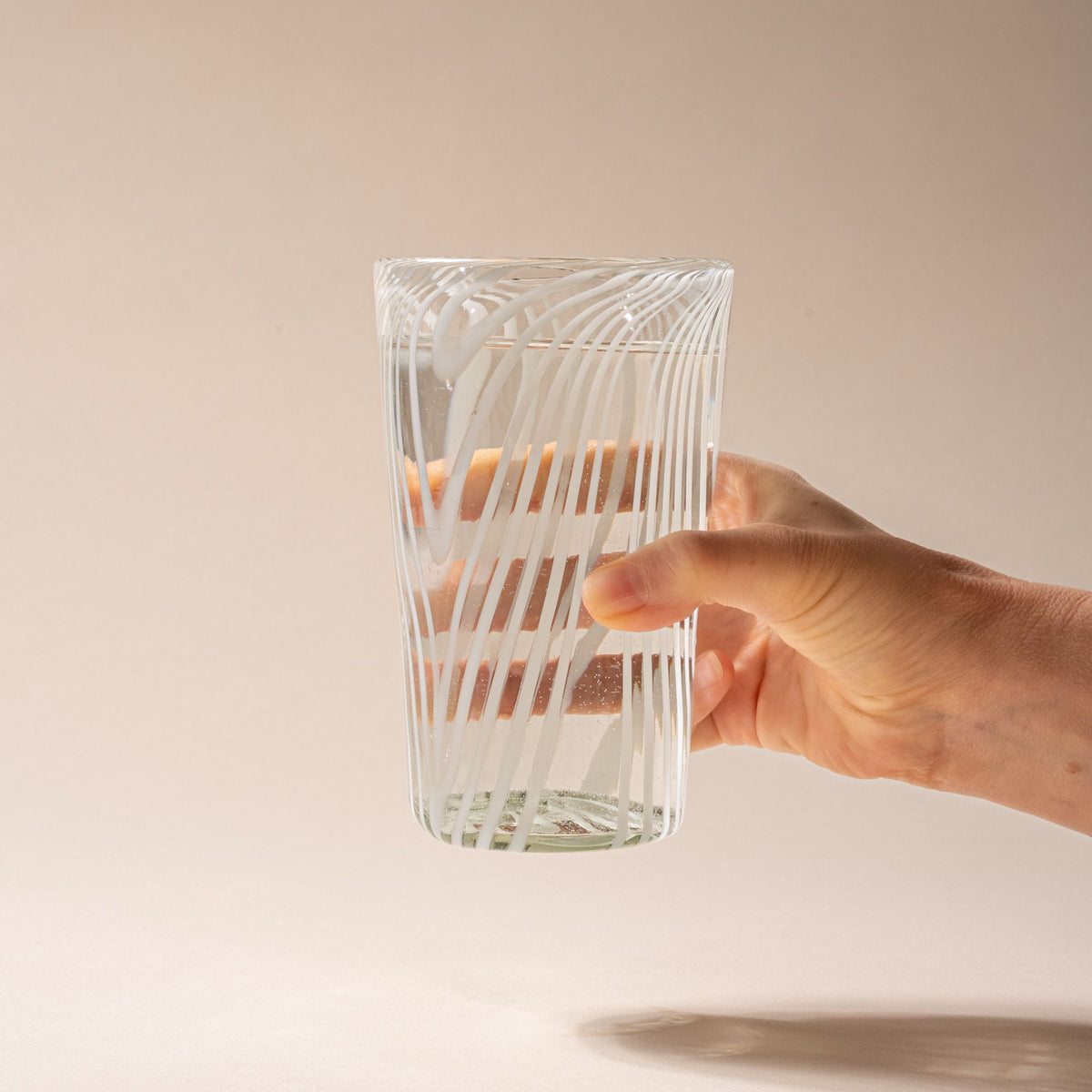 A hand holds out a clear pint glass with white diagonal swirl pattern on a neutral background.