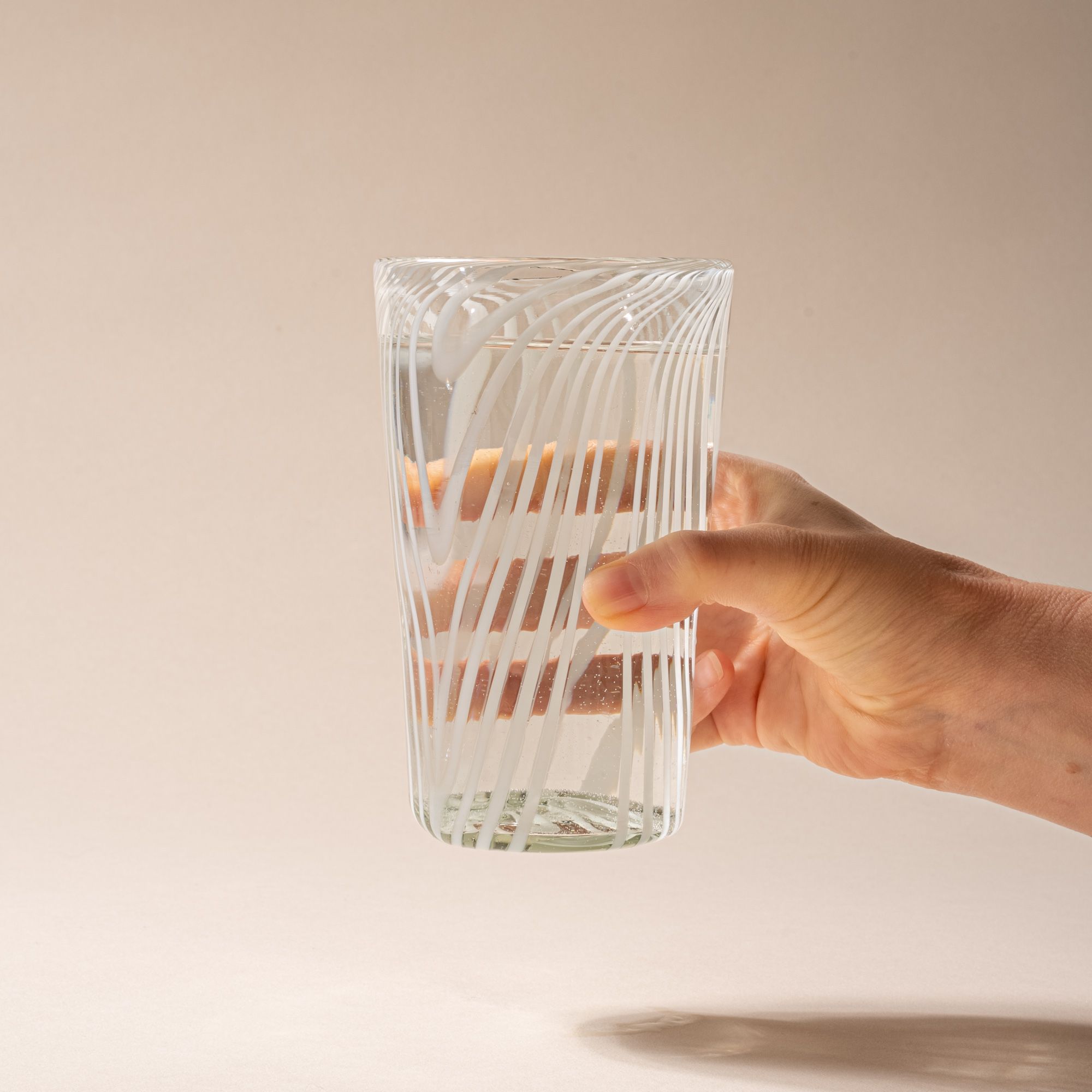 A hand holds out a clear pint glass with white diagonal swirl pattern on a neutral background.