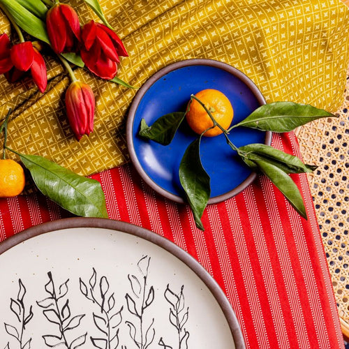 A small dessert-sized plate in a cobalt blue color with a plate with with a black drawing on it, both on red and yellow fabrics with oranges and flowers.