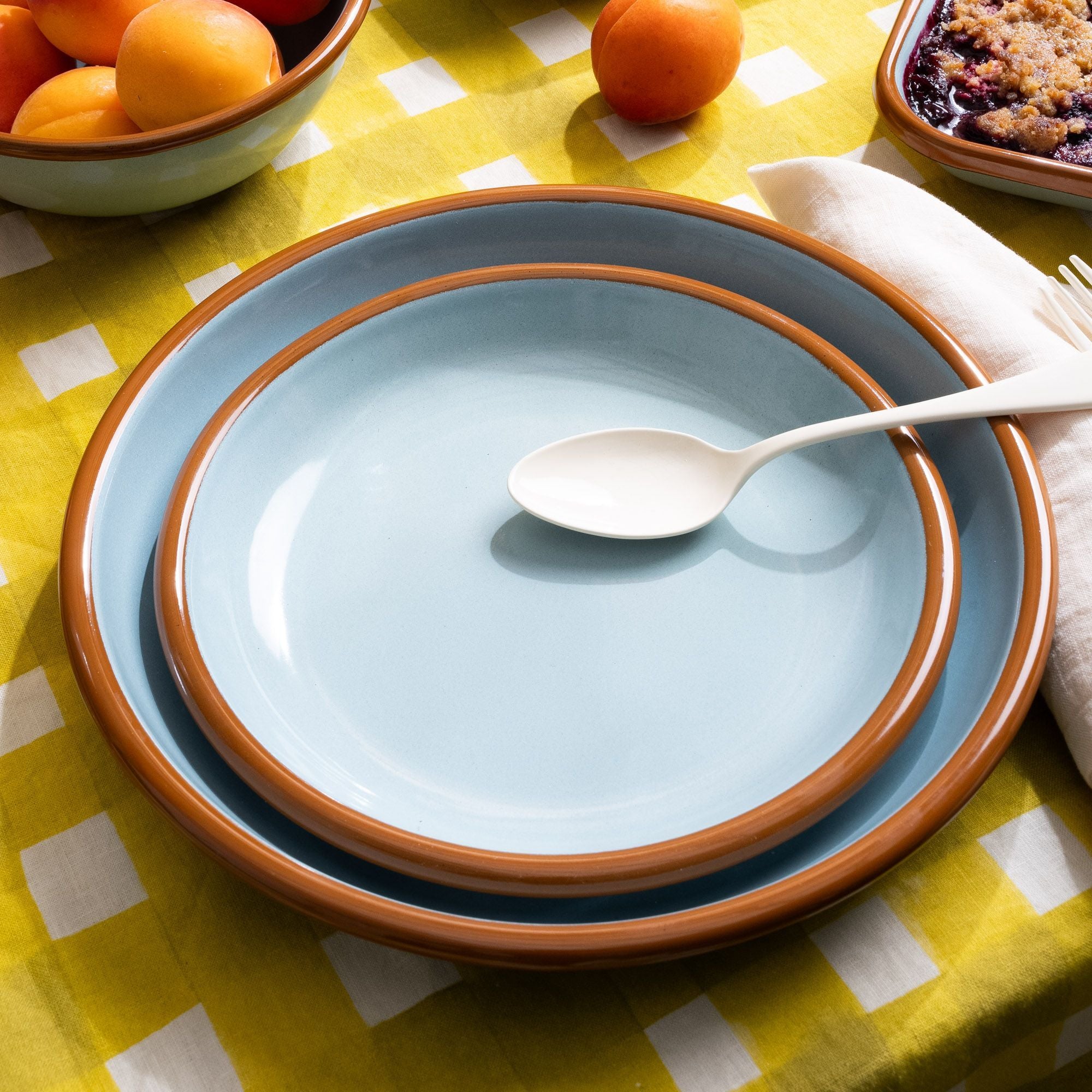 A nestled set of enamel side plate and dinner plate in robin's egg blue on a yellow patterned tablecloth with an enamel spoon.