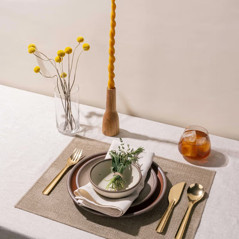 A table setting featuring a stack of ceramic plates, brass flatware, drink, taper candle, and more.