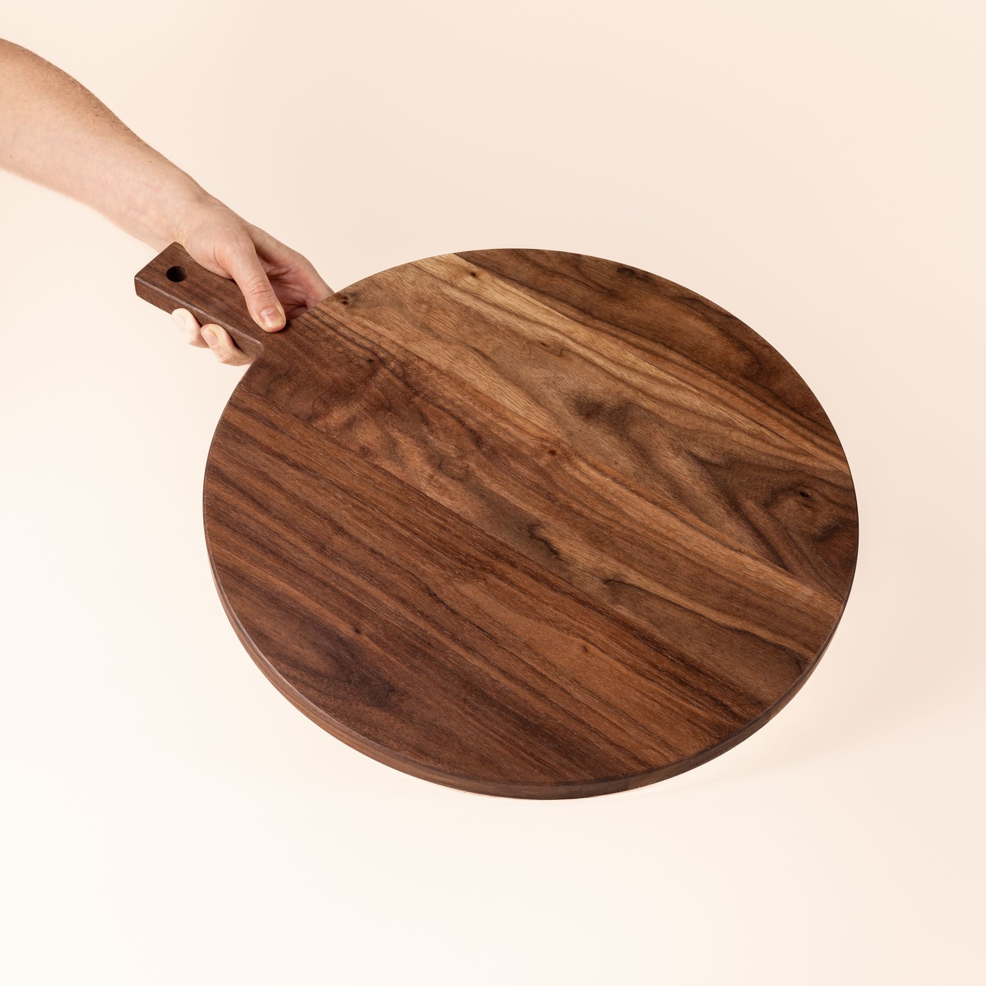 Hand holding a round wooden serving board with a short rectangular handle with a hole for hanging