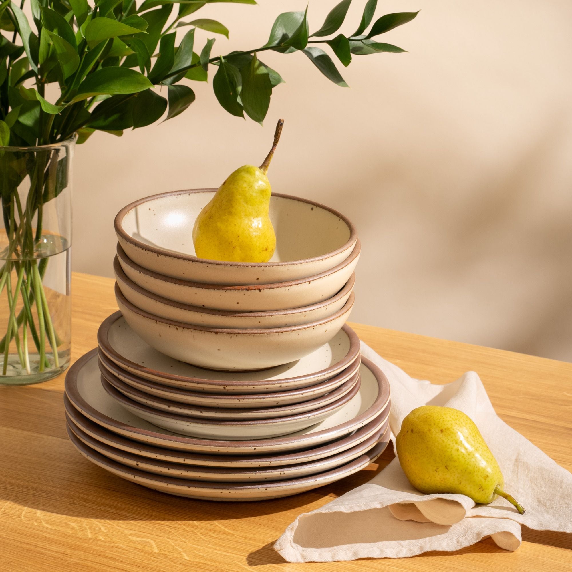 4 ceramic dinner plates, 4 side plates, and 4 shallow dinner bowls in a blue-grey color with iron speckles and unglazed rims are stacked all together on a countertop with a napkin and plant
