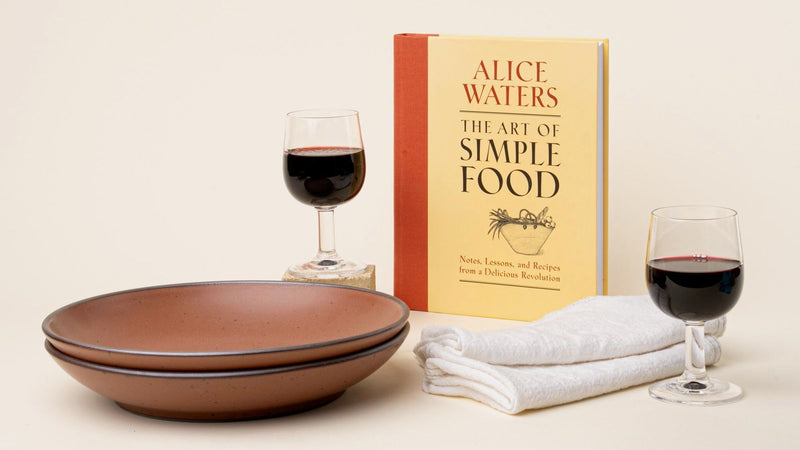 A copy of The Art of Simple Food by Alice Waters sits beside two glasses of red wine, stacked plates, and folded white napkins.