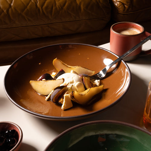Glossy brown ceramic bowl with sliced pears, figs, and cream, placed on a table beside a coral mug.