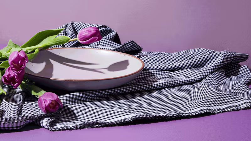 An East Fork Coupe plate in a pastel purple color with flowers and a background of purple hues, with a gingham black and white napkin.