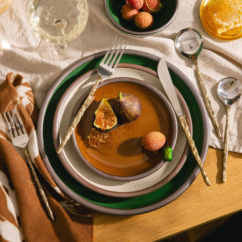 Layered caramel brown, white, and emerald green plates in earthy tones with figs, lychee, and honey, styled with golden flatware on a natural linen table setting.
