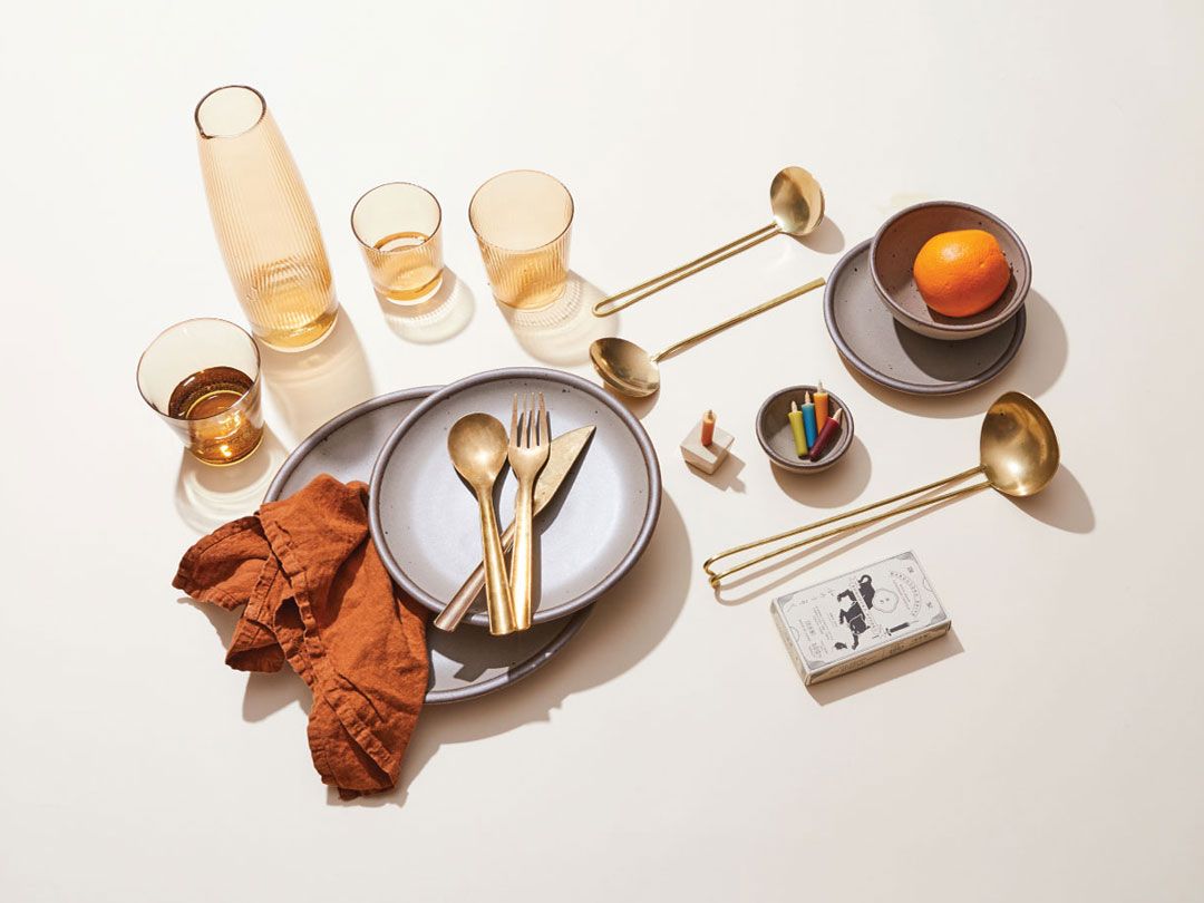 Ceramic plates and bowls in a soft gray glaze are paired with brass utensils and amber glassware on a light background. An orange, colorful candles in a small dish, and a rust-colored napkin add warmth to the arrangement.