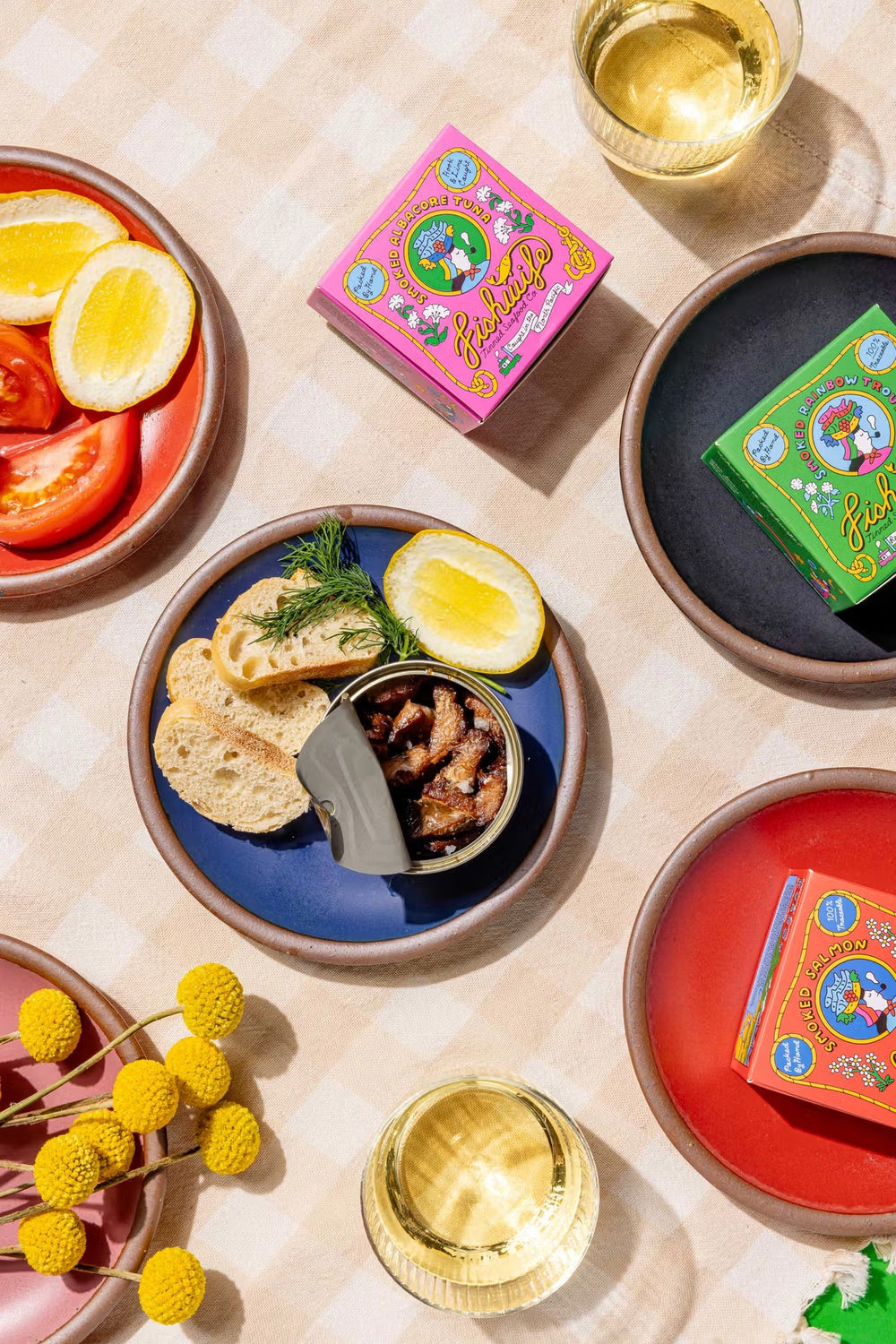 A vibrant picnic spread on a checkered cloth featuring open cans of fish, slices of bread, half lemons, and tomatoes on colored ceramic plates. Bright packaging and glasses of white wine accompany the arranged dishes, accented by fresh dill and yellow flowers.