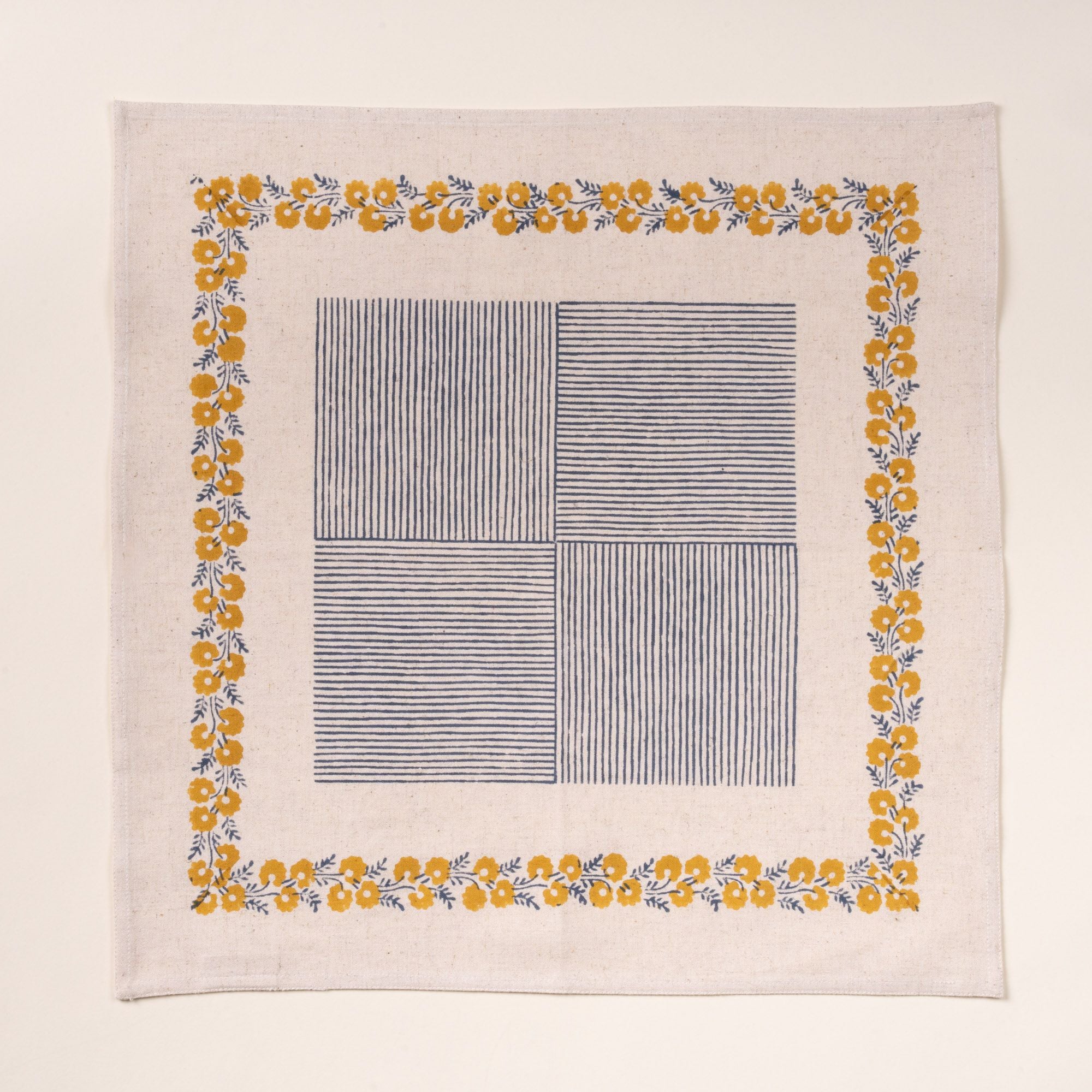 A cream-colored napkin with a delicate yellow floral border framing a central blue striped patchwork design, laid flat against a neutral background.