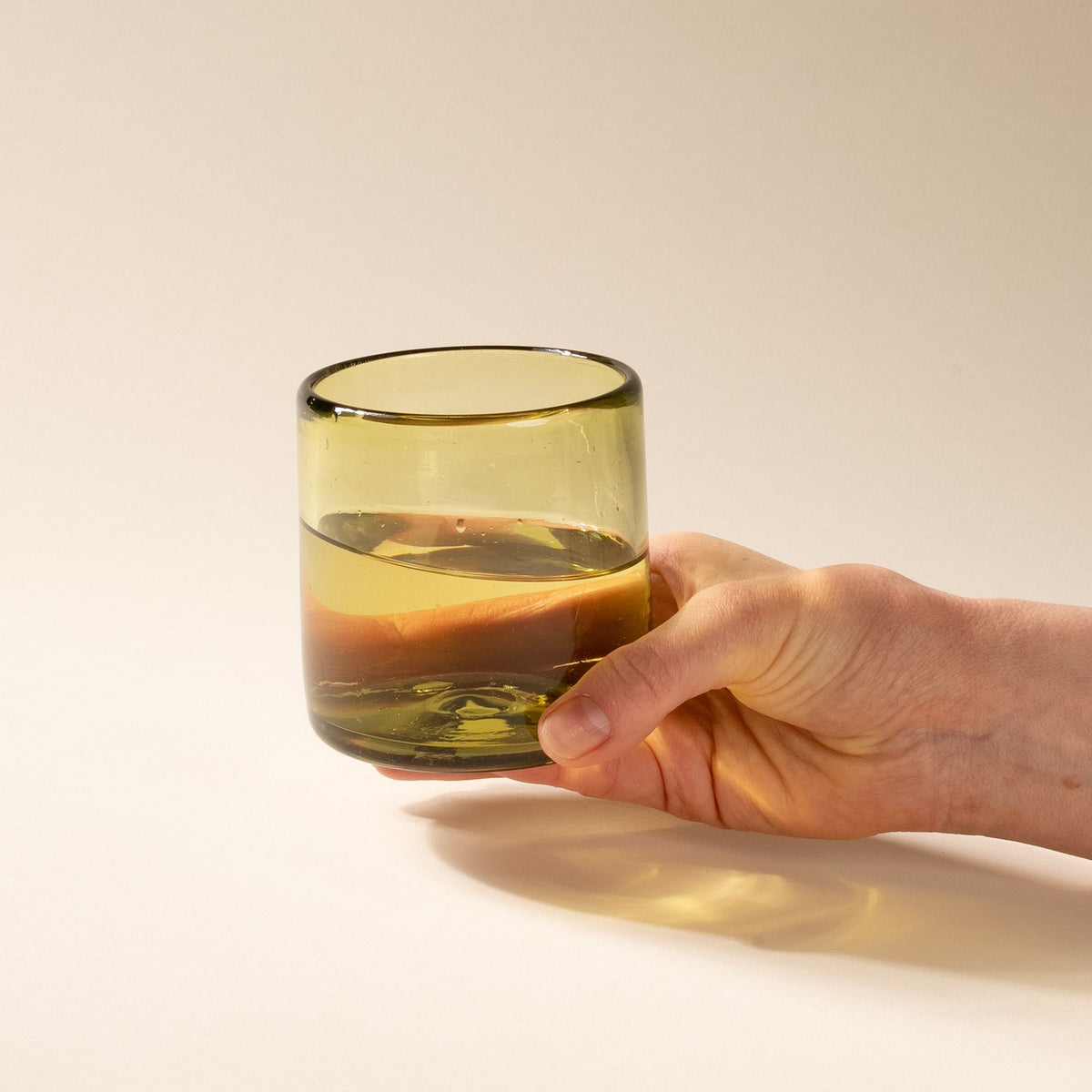 A hand holds an olive green short transparent drinking glass, filled with water.