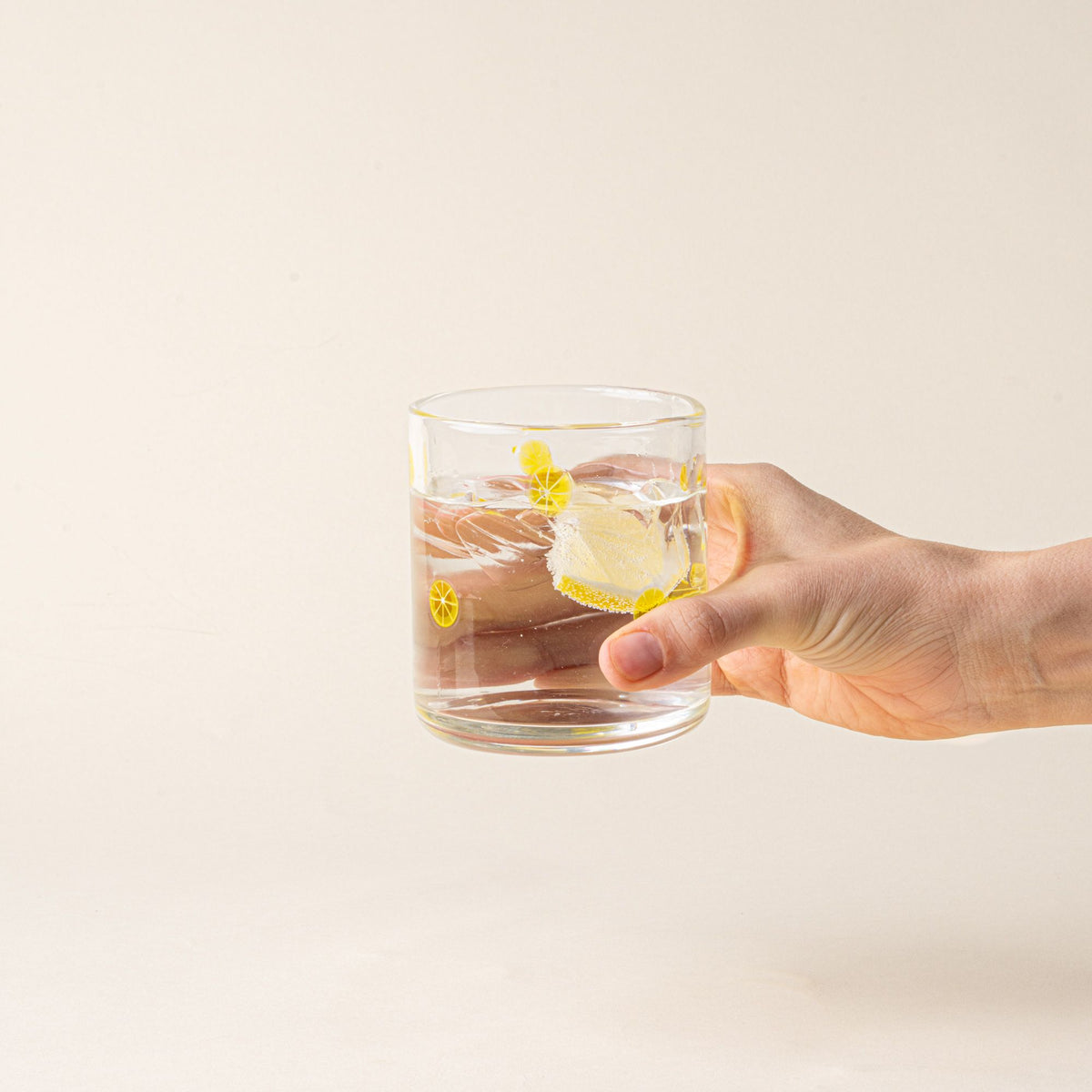 A hand holds a clear glass tumbler with yellow lemon slice motif, filled with water and a lemon slice.