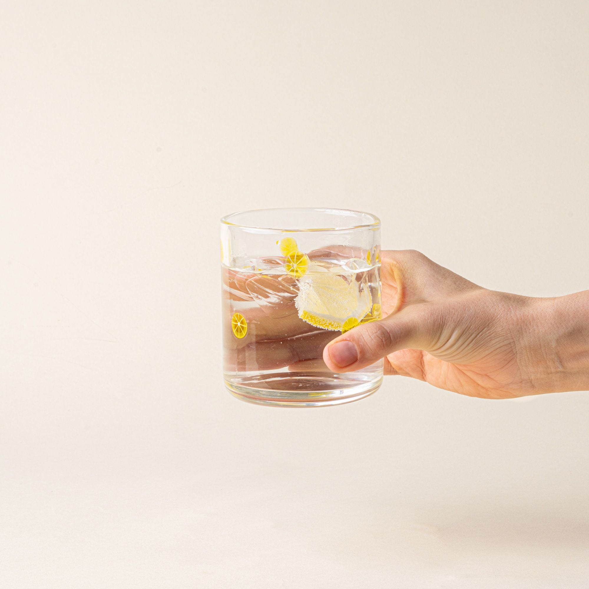 A hand holds a clear glass tumbler with yellow lemon slice motif, filled with water and a lemon slice.