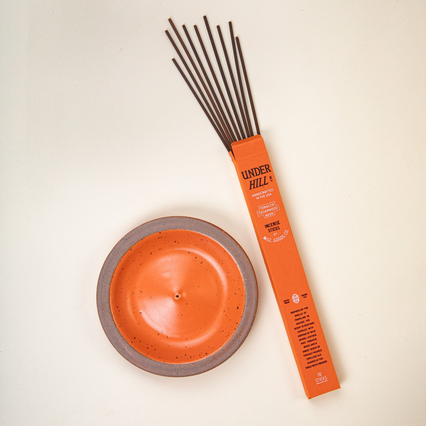 A ceramic plate-shaped incense stick holder with an unglazed rim and glazed in a bold orange color featuring lots of iron speckles. Beside the holder is a rectangular box of incense sticks coming out of the top.