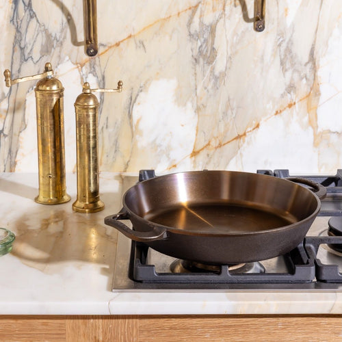 Cast iron skillet on gas stove with marble backsplash and brass salt and pepper mills in the background.