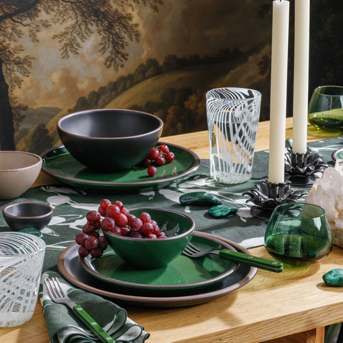 Table set with glossy emerald green ceramic plates and bowls, grapes, striped glassware, and tall white candles on a patterned table runner.