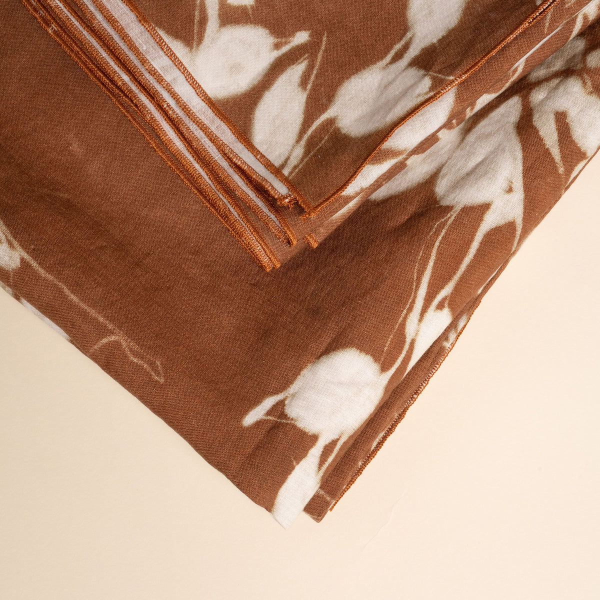 A closeup of a folded tablecloth in a brown color with white botanical print on a neutral background.