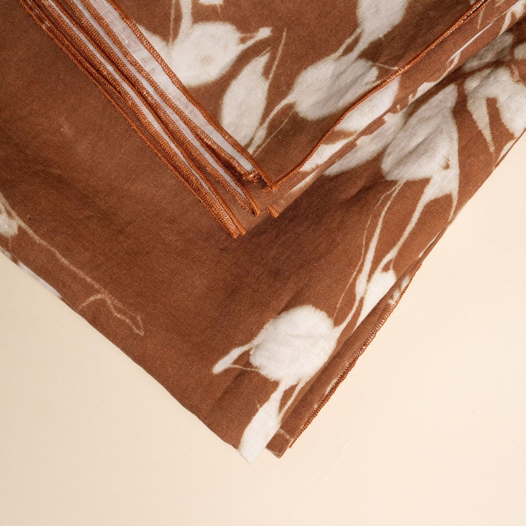 A closeup of a folded tablecloth in a brown color with white botanical print on a neutral background.