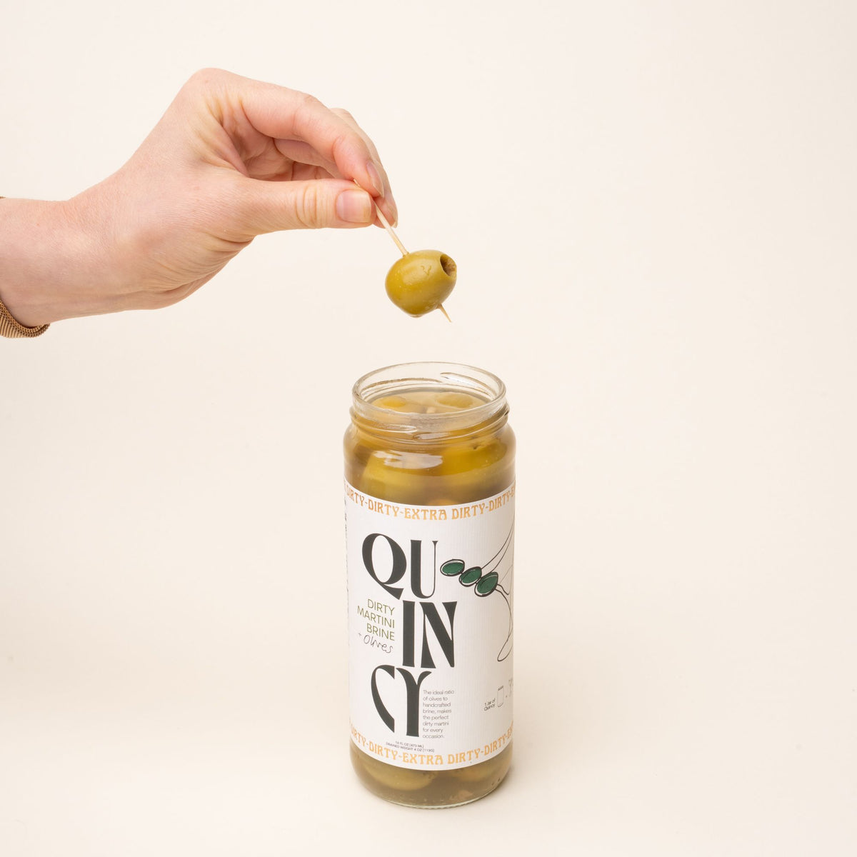 A hand holding an olive on a toothpick above a jar of Quincy Dirty Martini Brine with olives, featuring a white label with bold black text and olive illustrations.
