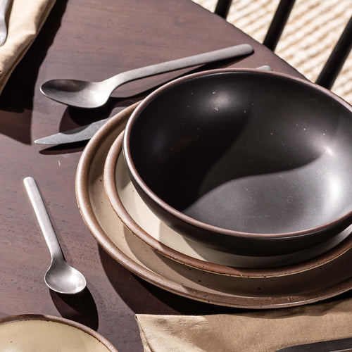 A place setting featuring a dinner plate, side plate, and shallow dinner bowl in graphite and neutral colors with steel flatware.