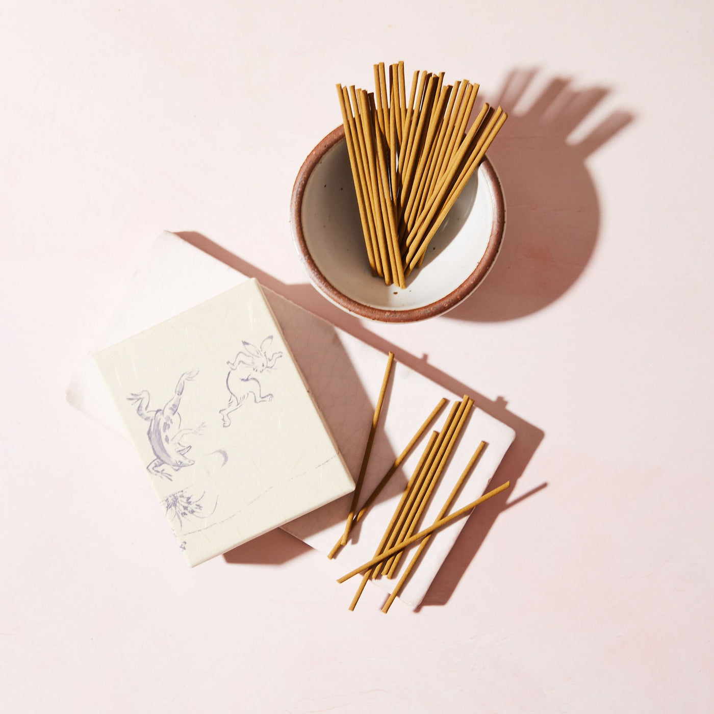 A small bowl filled with thin, brown incense sticks set on a light pink background. Next to the bowl is a decorative box with illustrations, and several incense sticks are laid beside it.