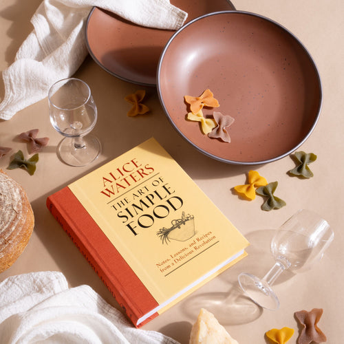 A copy of The Art of Simple Food lies beside speckled cool terracotta ceramic plate bowls, colorful pasta, wine glasses, and white napkins on a tan surface.