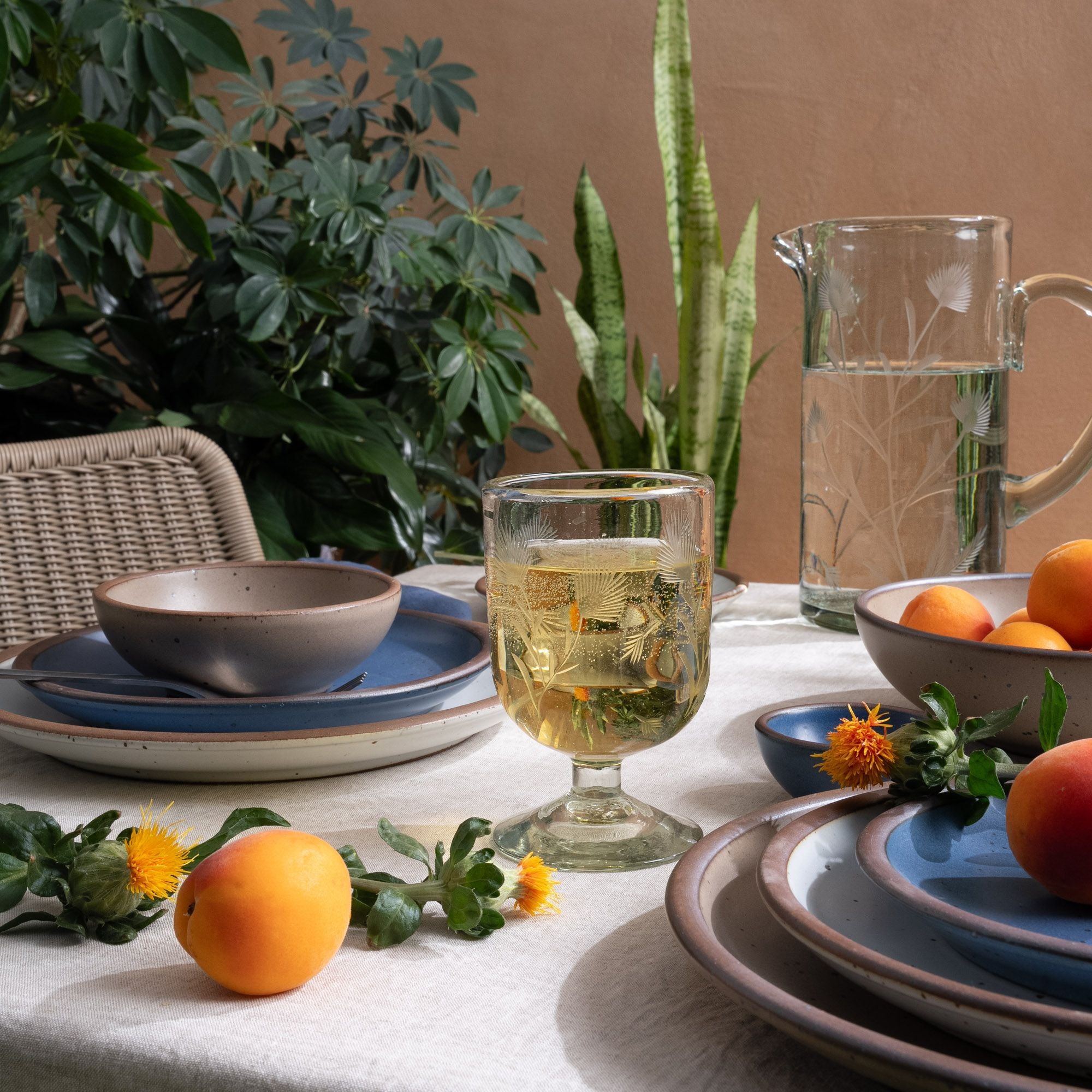 A clear short wine glass made of recycled glass with etched thistle designs on it, filled with white wine, on a table with ceramic dinnerware, plants, fruit and flowers.