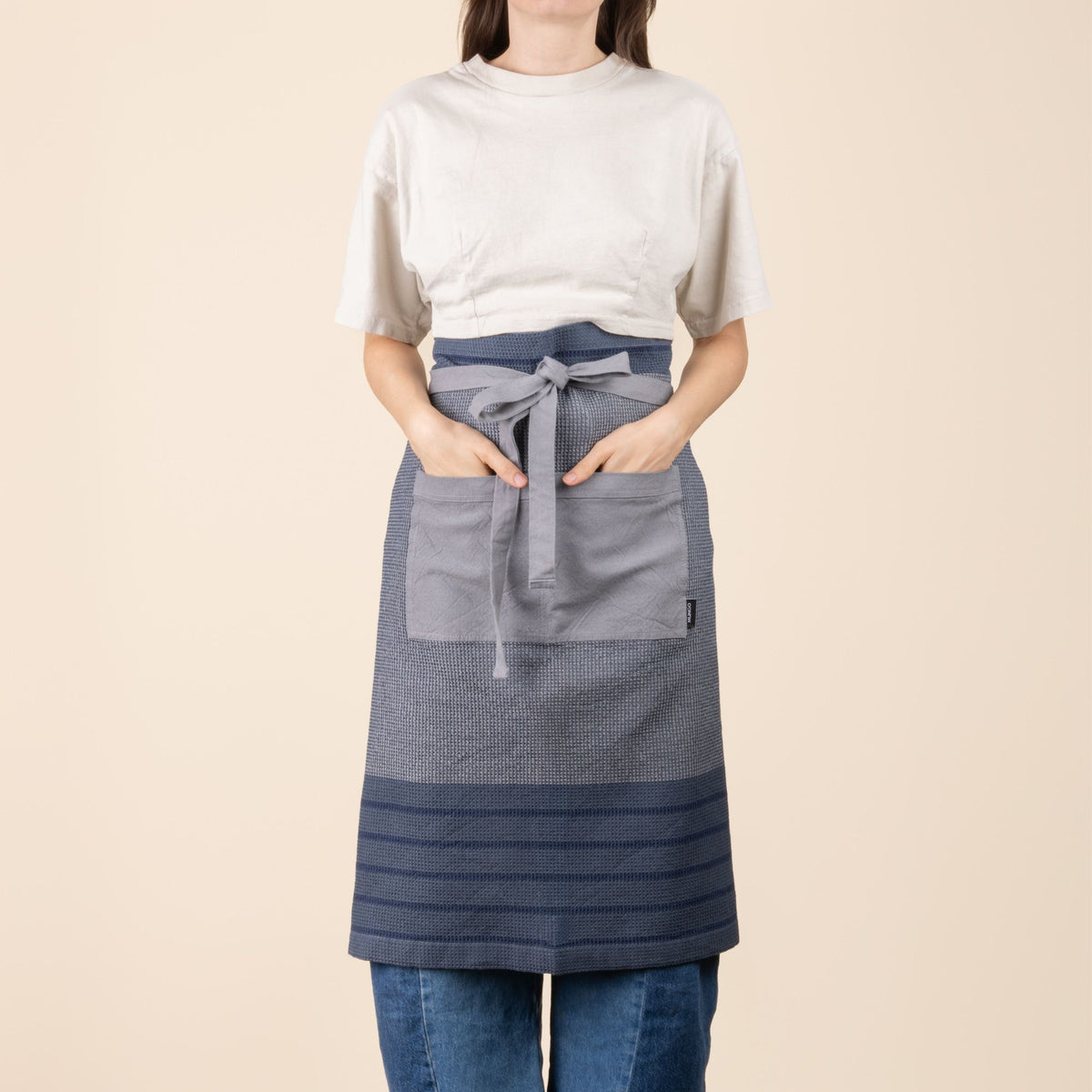 A person wears a woven kitchen waist apron in navy and slate blue with light blue and rust stripe accents with a front pocket and long tie straps.