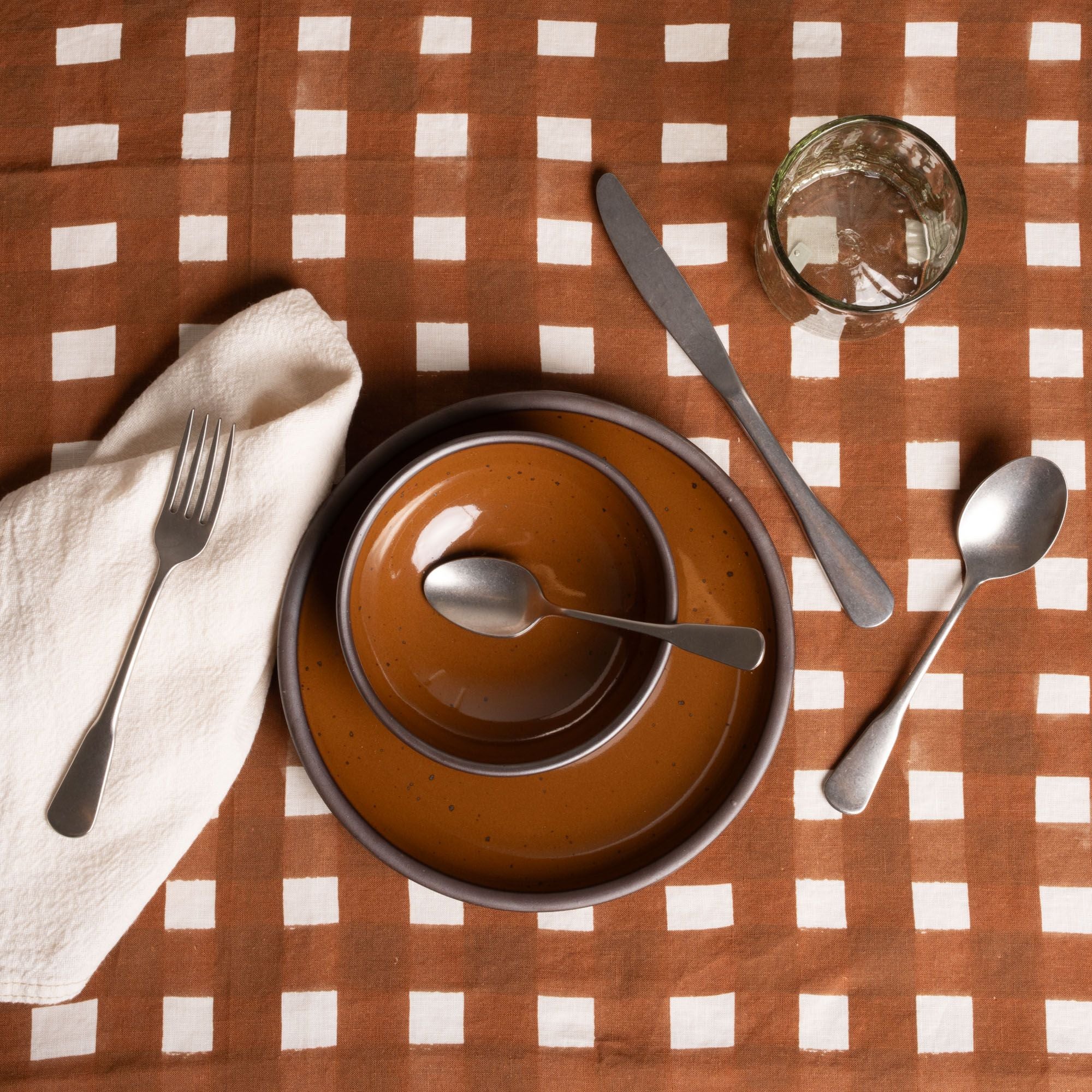 Place setting on brown and white crosshatch tablecloth with stacked brown ceramic plates, utensils, glass, and napkin.