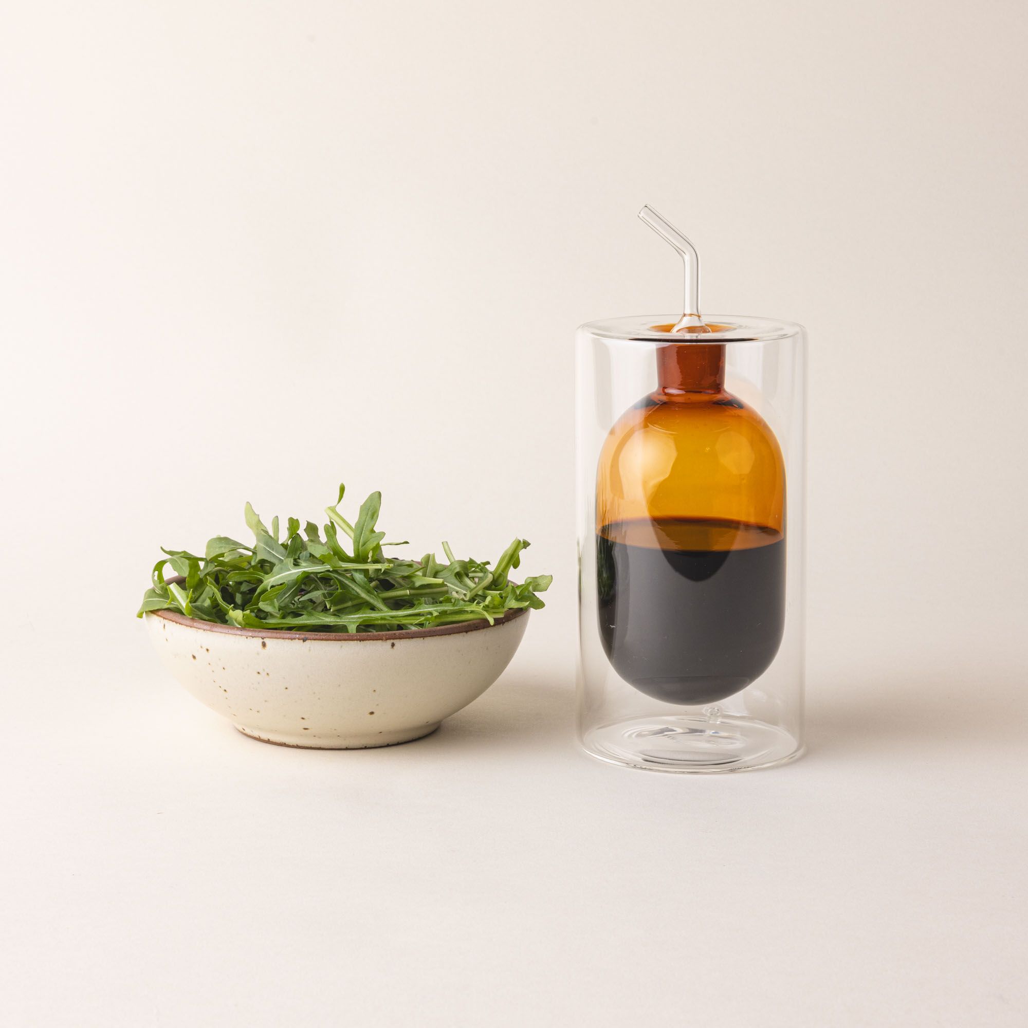 An artful glass cruet in an orange color and is next to a small side salad. There is a rounded bulb that acts as a container for the cruet, surrounded by a doubled glass wall. The cruet is filled with oil.
