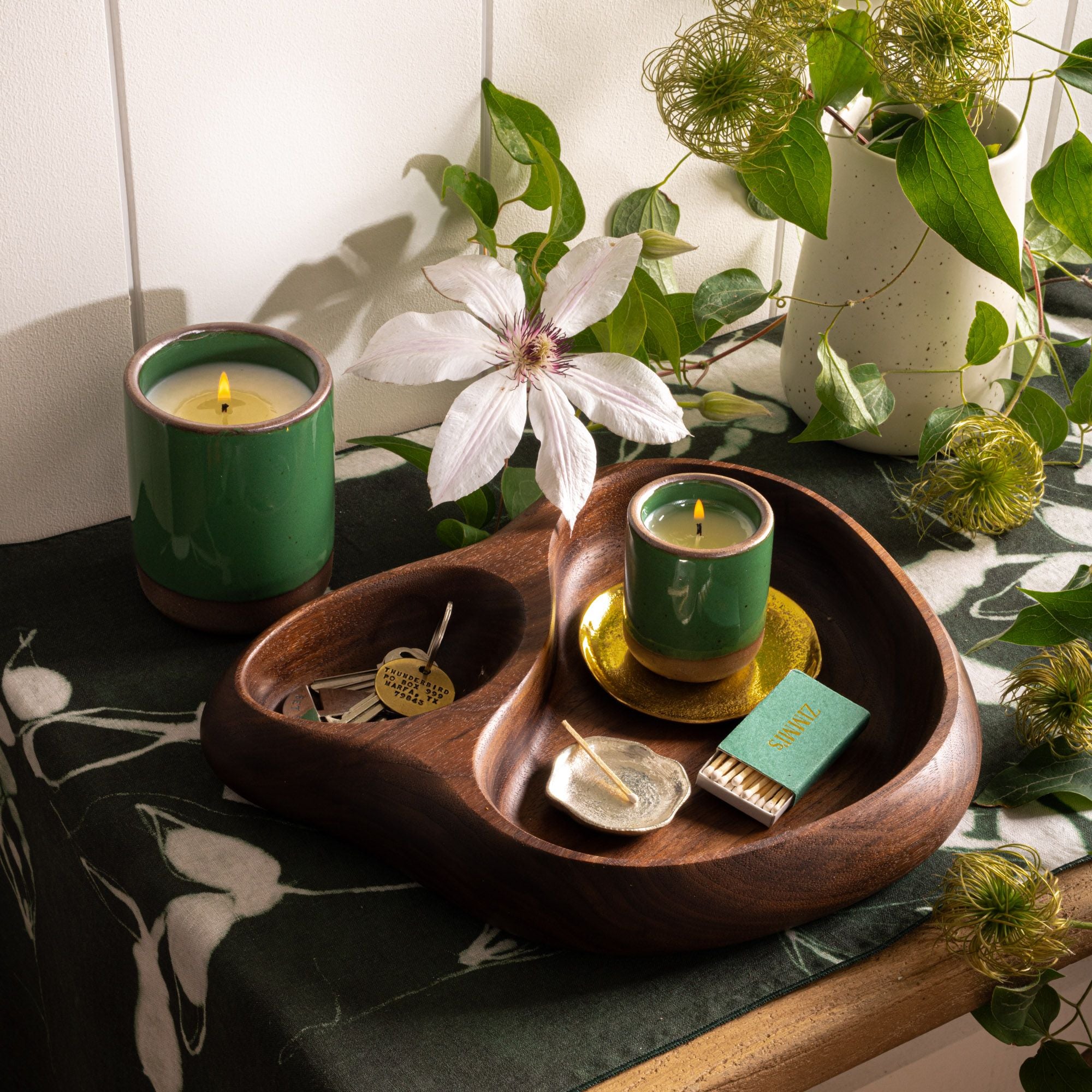 Two glossy green ceramic candles in a sculptural wooden tray with keys, matches, and a flower on a patterned fabric.