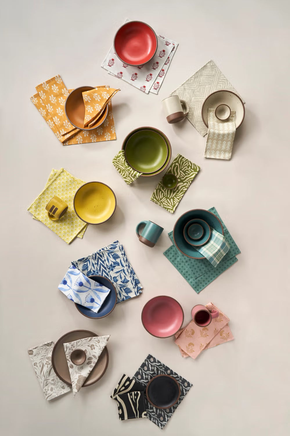 A variety of colorful ceramic dishes, bowls, and mugs arranged on patterned napkins, displayed on a neutral background.