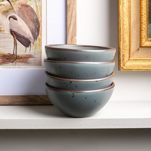 On a shelf in front of framed art pieces, sits a stack of 4 small dessert sized rounded ceramic bowls in a blue-grey color featuring iron speckles and an unglazed rim