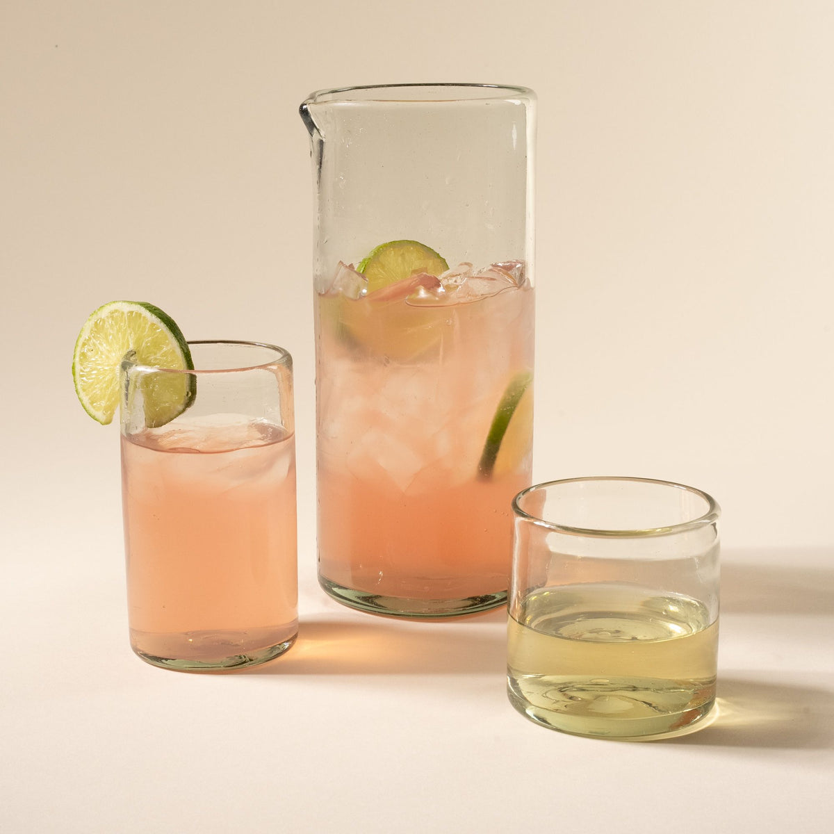 A tall tumbler, large pitcher, and short tumbler, of clear glassware all filled with drinks.