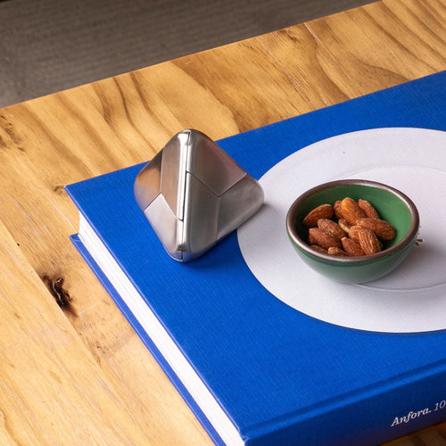 Silver geometric pyramid sculpture next to a small green bowl of almonds on a large blue art book on a wooden table.