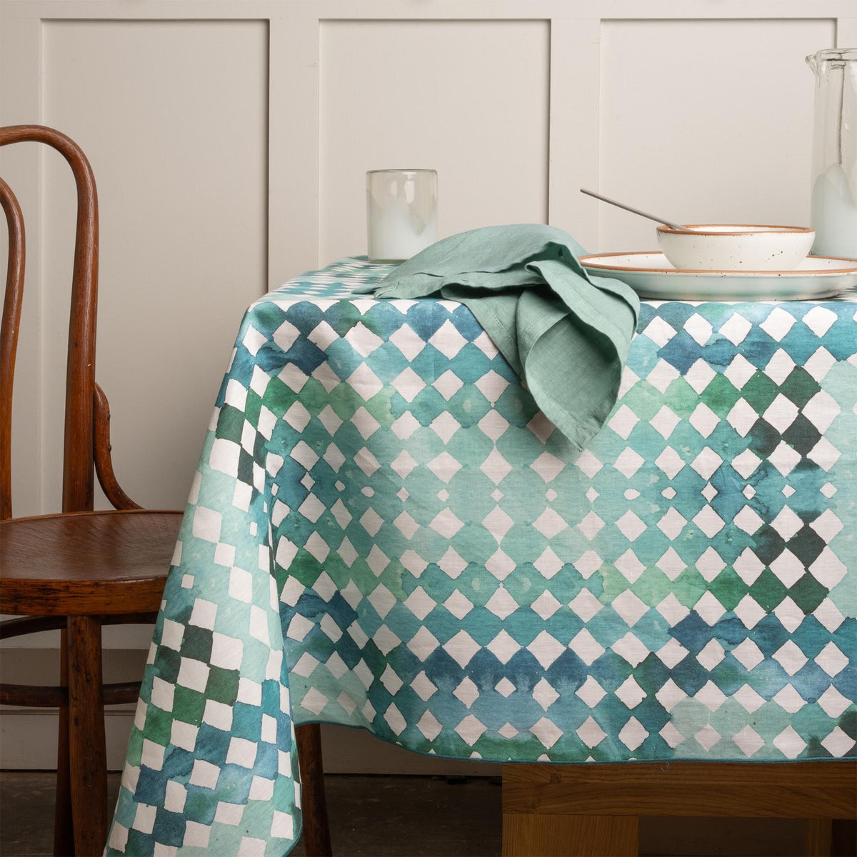 Table set with white ceramic dishware on a teal watercolor diamond patterned tablecloth, with a wooden chair and linen napkin nearby.