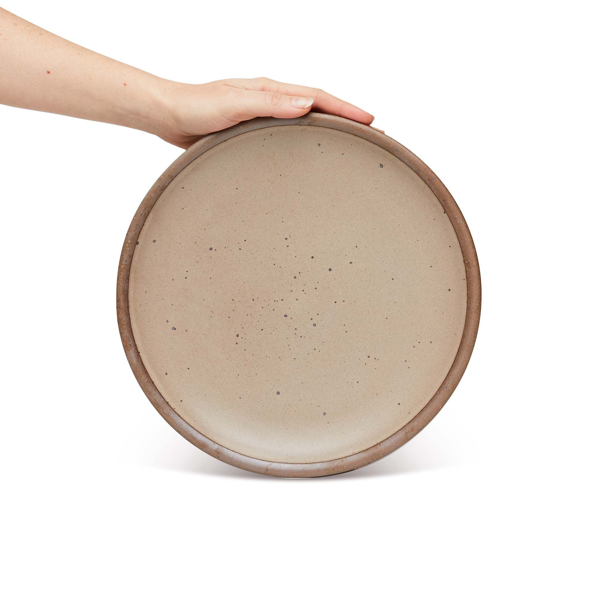 Hand holding a dinner sized ceramic plate in a warm pale brown color featuring iron speckles and an unglazed rim.